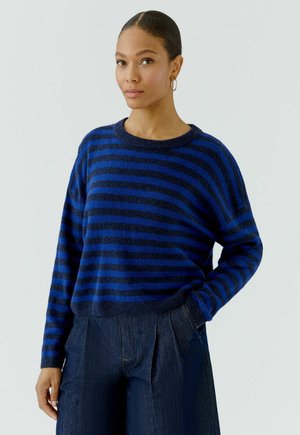Vrouw met haar naar achteren gebonden, die gouden oorringen draagt, een blauw en marineblauw gestreepte trui en donkerblauwe wijde broek.
