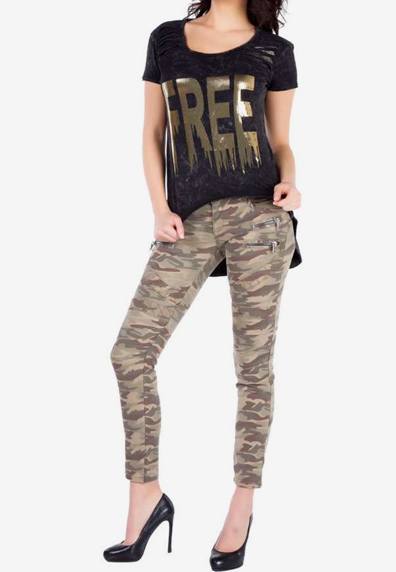 Femme portant un T-shirt noir usé avec le texte doré "FREE", pantalon camouflage avec détails zippés, et chaussures à talons noires.