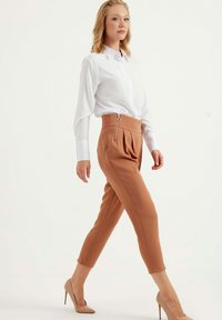 Beige hoge taille broek met zijplooien en een voorzijde ritssluiting, gecombineerd met een witte blouse met kraag en nude hoge hakken.