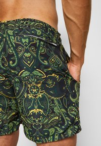 Schwarze Badehosen mit grünem und gelbem Paisley-Muster, elastischem Bund und seitlichen Taschen. Hergestellt aus leichtem, schnell trocknendem Material.