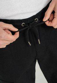 Schwarze Jogginghose mit strukturierter Oberfläche, elastischem Bund und Metallösen. Eine Hand greift an den geflochtenen Kordelzug vorne.