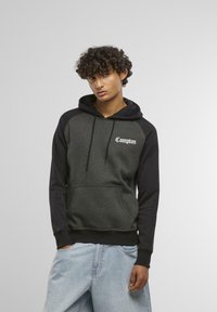 Sweatshirt à capuche avec un corps gris foncé et des manches noires, comportant une poche avant et un logo "Compton" en cursive blanche. Texture douce.