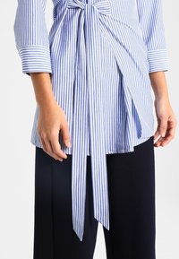 Bluzka w niebiesko-białe paski z detalem w postaci związania w talii, z rękawami do trzech czwartych i sylwetką w kształcie litery A, zestawiona z ciemnymi spodniami.