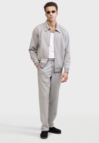 Chaqueta gris con cremallera y cuello y puños de canalé, combinada con pantalones de chándal a juego con cordón. El modelo lleva gafas de sol y zapatos negro deslustrados.