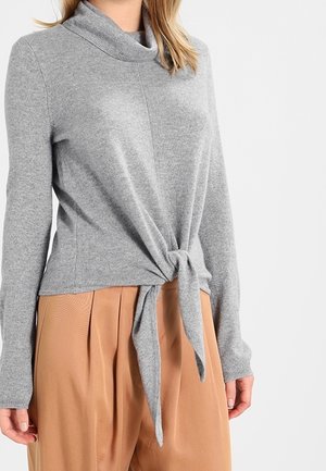 Femme portant un pull à manches longues gris clair avec un détail noué à l'avant, assorti à un pantalon plissé taille haute de couleur camel.