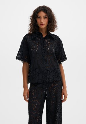 Kobieta o kręconych włosach, ubrana w czarną koszulę na guziki oraz spodnie z kwiatowym ażurowym wzorem, stojąca na tle jednolitego jasnego tła.