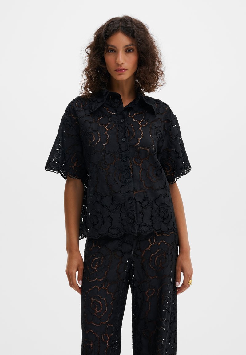 Γυναίκα με σγουρά μαλλιά που φοράει μαύρο πουκάμισο με κουμπιά και παντελόνι με λουλουδάτο δαντελωτό μοτίβο, στέκεται μπροστά σε απλό ανοιχτό φόντο.