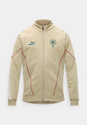 Jachetă sport bej cu fermoar, tiv roșu, logo Puma verde, text „KING” pe pieptul stâng și emblemă circulară a echipei pe pieptul drept.