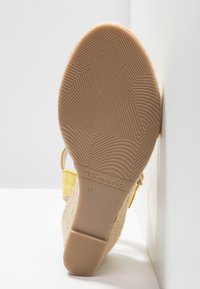 Semelle de sandale compensée beige avec une semelle extérieure géométrique texturée, taille 37, et bride jaune, appuyée contre un mur blanc.