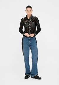 Camicetta in pizzo nero con tasche in denim, chiusura a bottoni e maniche lunghe, abbinata a jeans svasati blu e mocassini neri, su sfondo bianco.