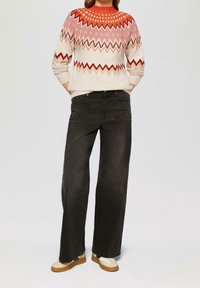 Lichtgekleurde gebreide trui met rode en roze geometrische patronen, gecombineerd met wijde zwarte jeans en bicolor schoenen.