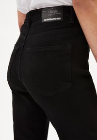 Persona que lleva vaqueros de mezclilla negros con un bolsillo trasero visible y etiqueta de marca ARMEDANGELS en la cintura.