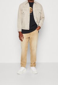 Beige jeansjacka över en svart T-shirt med en mån-grafik, tillsammans med ljusbeige jeans och vita sneakers. En enkel, avslappnad look.