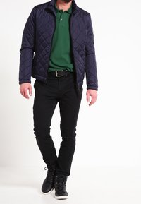 Giacca trapuntata blu navy con colletto alto, polo verde, pantaloni neri e sneakers nere. Presenta cerniere e una texture liscia.