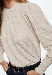 Beige tekstureret bluse med høj krave, samlet detaljering og lange ærmer, parret med et mørkt denimbælte og jeans.