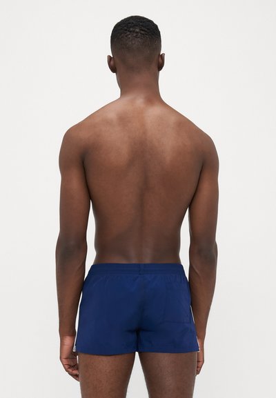 Shorts de bain bleu marine avec une taille élastique, poche arrière et tissu lisse, vus de dos sur une personne.