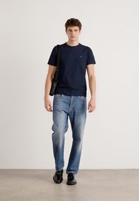 Un tricou navy cu maneci scurte, blugi albaștri lejeri și pantofi negri. Modelul poartă o geantă de umăr neagră. Design simplu, croi casual, textură netedă.