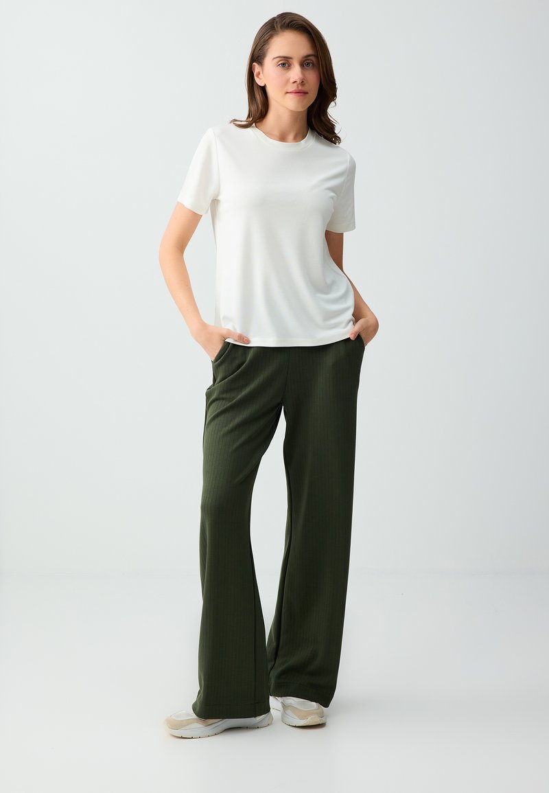Jeune femme debout, les mains dans les poches, portant une chemise blanche à manches courtes, un pantalon large vert foncé et des baskets blanches sur un fond uni.