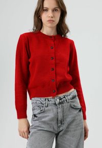 Cardigan rouge et duveteux avec un col rond, une fermeture à boutons et des manches longues. Associé à un jean en denim gris clair à taille haute.