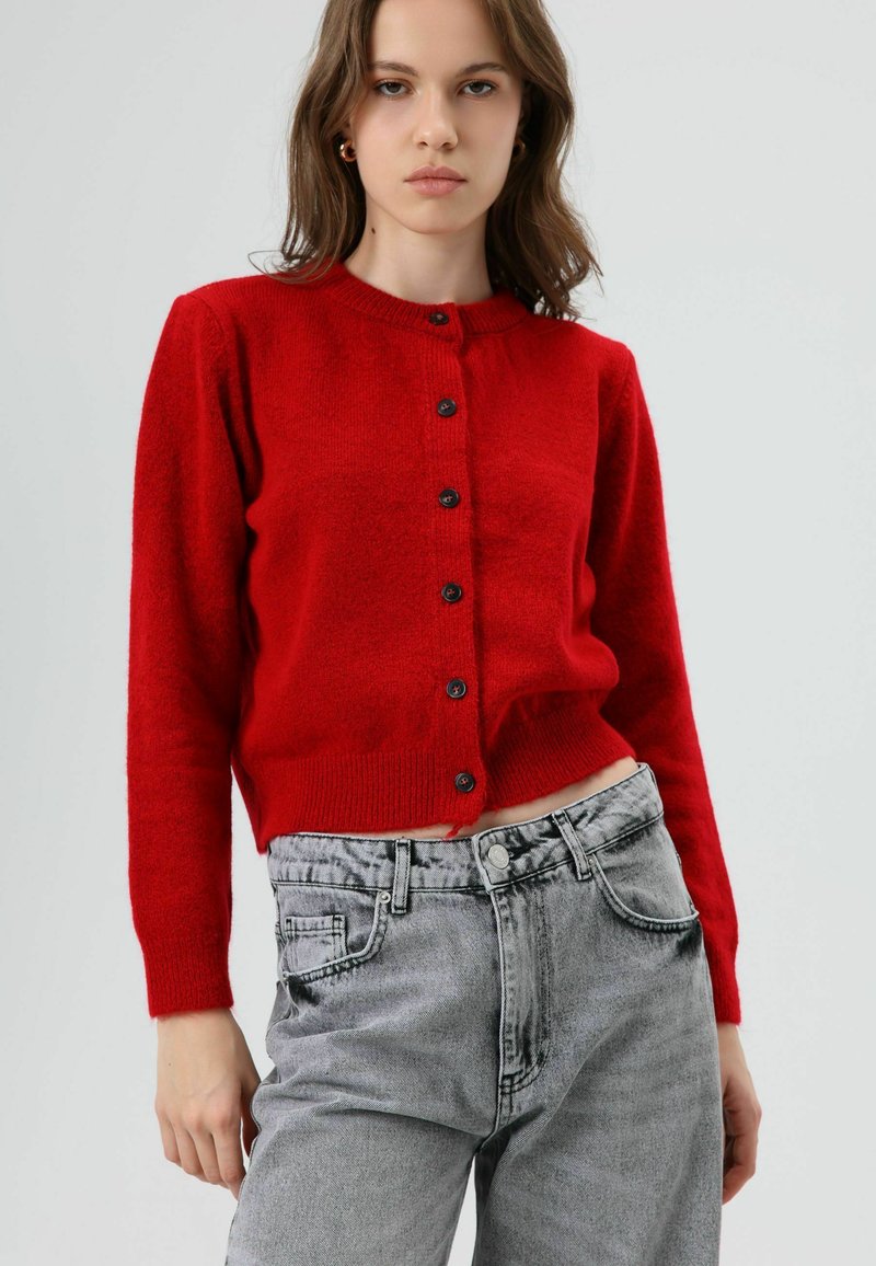 Cardigan rouge et duveteux avec un col rond, une fermeture à boutons et des manches longues. Associé à un jean en denim gris clair à taille haute.