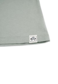 Helle grüne Baumwoll-T-Shirt mit einem gesäumten Ärmel und einem gewebten Etikett mit "B&R" und gekreuzten Utensilien in Schwarz. Einfaches Design, keine Muster.