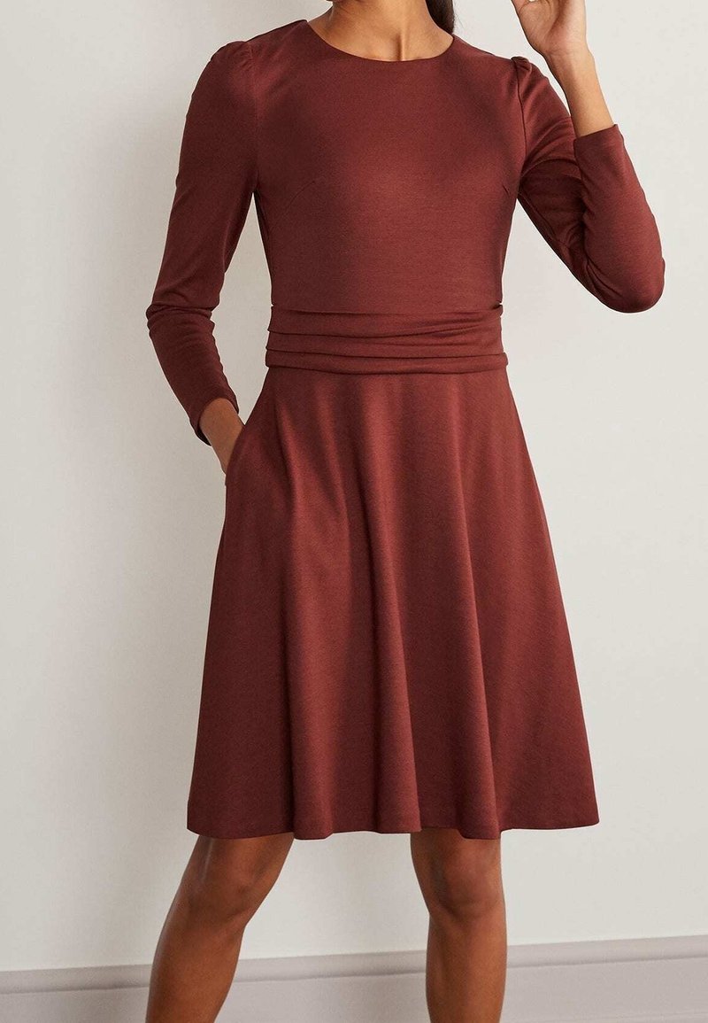Femme portant une robe marron rouille à manches longues, longueur jusqu'aux genoux, avec un corsage ajusté et une ceinture plissée, debout contre un mur uni.