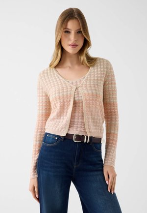 Młoda kobieta nosząca jasnoróżowy sweter na drutach, do tego dopasowany top i ciemnoniebieskie dżinsy, stojąca na tle jednolitego, jasnego tła.