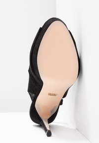 Chaussure à talon haut en daim noir avec bout pointu vue de la semelle, appuyée contre un coin blanc du mur et du sol.