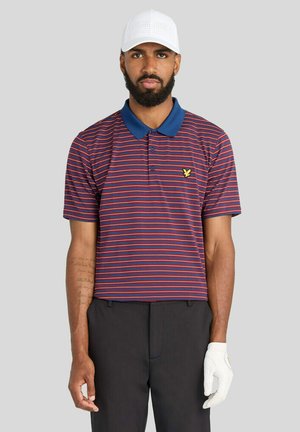 Uomo con berretto bianco, polo a righe rosse e blu, pantaloni neri e guanto da golf bianco, in piedi davanti a uno sfondo grigio uniforme.