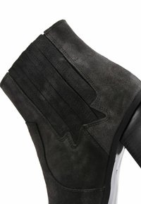 Black suede ankle boot featuring a ribbed panel and textured black sole. The design showcases smooth and slightly matte finishes.