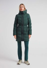 Abrigo acolchado verde con un alto cuello, cintura ceñida, textura acolchada y detalles en negro en la cremallera, combinado con pantalones verdes y calzado ligero.