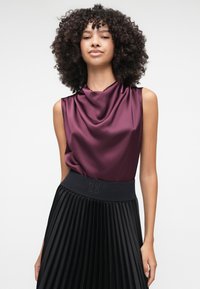 Top senza maniche in satin bordeaux con scollatura drappeggiata. Abbinato a una gonna midi plissettata nera, con una vita decorata da logo in rilievo.