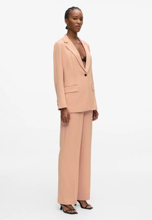 Femme portant un blazer oversize pêche et un pantalon large assorti, un soutien-gorge noir et des sandales à talons noires, debout devant un fond blanc.