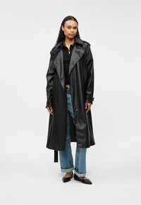 Czarny, błyszczący, długi trencz z szerokim kołnierzem i zapięciem na guziki, połączony z jasnoniebieskimi dżinsami i czarnymi szpiczastymi balerinami.