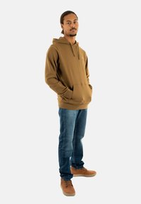 Felpa marrone con tasca frontale e cordino, abbinata a jeans blu e calzature marroni, caratterizzata da un design semplice e un colore uniforme.