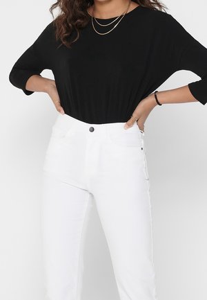 Femme portant un haut noir à manches longues rentré dans un jean blanc taille haute, mains posées sur les hanches, avec des colliers et bracelets dorés en couches.