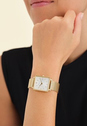 Reloj de pulsera de malla dorada con una esfera cuadrada blanca, marcadores de horas metálicos y una corona sencilla. Lleva puesto en una muñeca sobre un fondo negro.