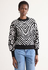 Fekete-fehér kötött pulóver, merész zebra mintával, puffos ujjakkal és ribbelt nyakkal. Világoskék farmerrel párosítva.
