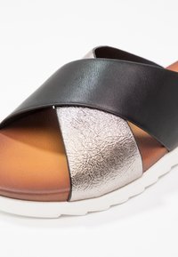 Schwarze und metallisch silberne Sandale mit Überkreuzriemen, strukturierter Oberseite, brauner Innensohle und weißer Sohle, mit einem runden Zehendesign.