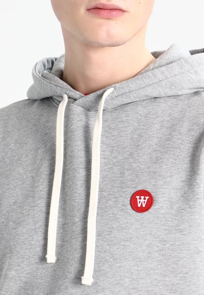 Šedá mikina s jemnou textúrou, ktorá má okrúhlu červenú nášivku s bielym logom a kontrastné biele šnúrky na kapucni.