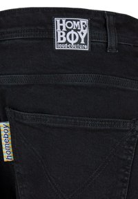Los pantalones vaqueros de mezclilla negra presentan una etiqueta visible con "HOME BOY loud-couture". Incluyen detalles de costura y una pequeña etiqueta multicolor en el costado.