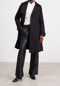 Svart trenchcoat med dubbelknäppt design, knappdetaljer och axelklaffar. Bärs över svarta läderbyxor och stövletter.