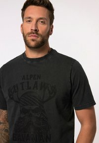 Sort t-shirt med korte ærmer og rund halsudskæring, med et stort grafisk design af et kranium med gevir og teksten "ALPEN OUTLAWS BAVARIAN."