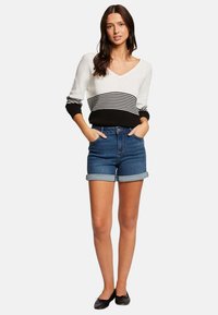 Maglione con scollo a V, bianco con strisce orizzontali nere e grigie; abbinato a pantaloni corti in denim blu arrotolati. Il modello sta in piedi con le mani nelle tasche.
