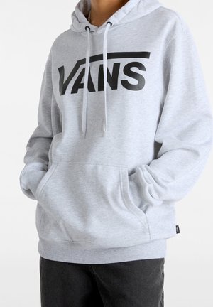 Persoon draagt een lichtgrijze Vans-hoodie met buidelzak en zwart Vans-logo, handen deels in de zakken, gecombineerd met een donkere broek.