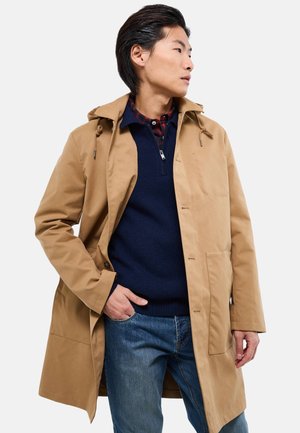 Homme portant un manteau à capuche beige sur un pull bleu marine à demi-fermeture éclair et une chemise à carreaux, posant avec une main dans la poche de son jean, regardant sur le côté.