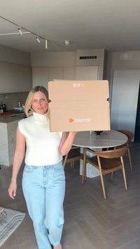 Brown Zalando box held in front of a person, with a white, ribbed, sleeveless top and light blue jeans, in a modern interior setting.