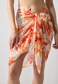 Blomstrete omslagsskjørt med et lyst oransje og gult mønster på lett, gjennomsiktig stoff, knyttet foran med en knyttet detalj.