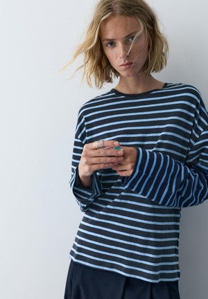 Ung kvinde med blondt hår iført blå stribet langærmet skjorte, holder hænderne nær brystet, foran en ensartet lysegrå baggrund.