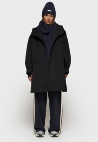 Zwarte waterdichte parka met verstelbare trekkoordkap en voorrits, gecombineerd met een marineblauwe sjaal, losse donkere broek en schoenen met zilveren accenten.